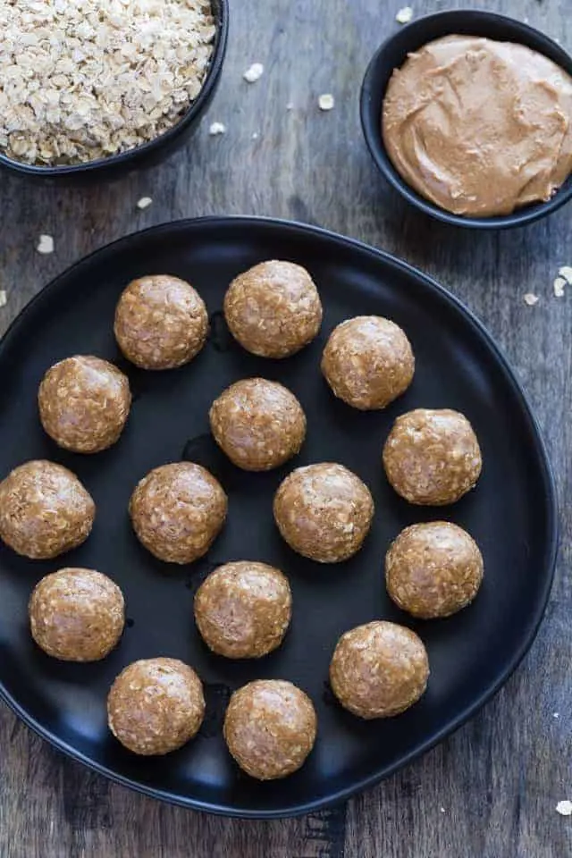 No Bake Almond Butter Balls neatly arranged on a black plate with a bowl of honey and a bowl of oats sitting behind the plate.