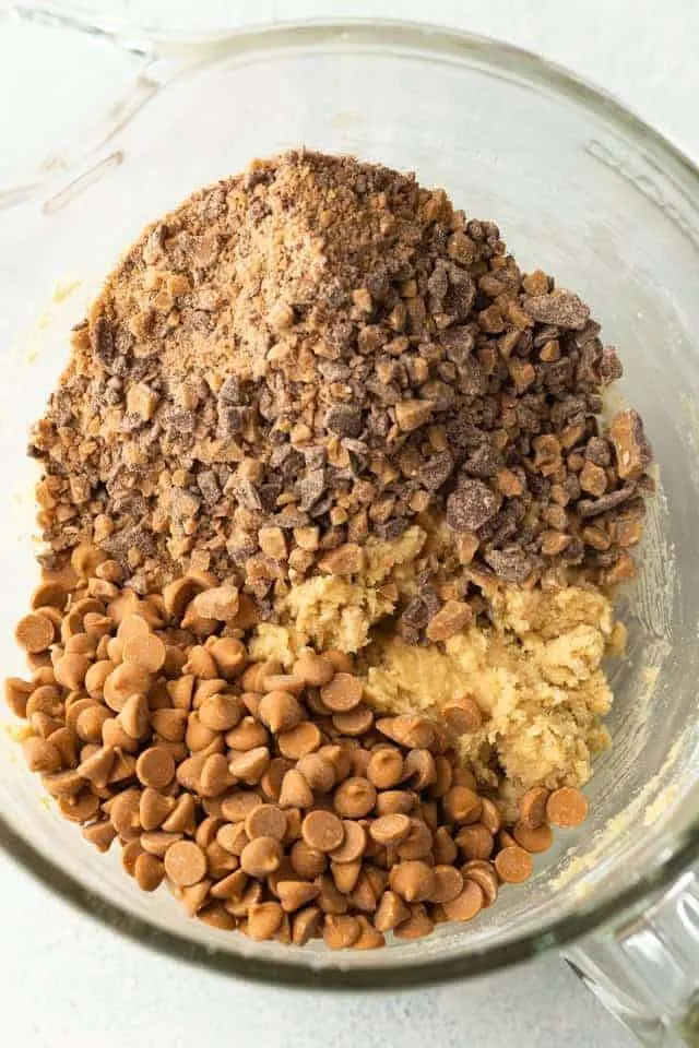 butterscotch chips and chocolate toffee pieces added to cookie dough in a glass bowl