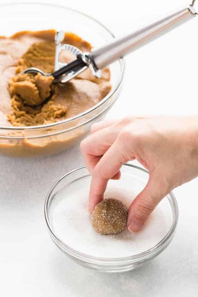 A hand rolling gingersnap cookie dough in white sugar in a glass bowl.
