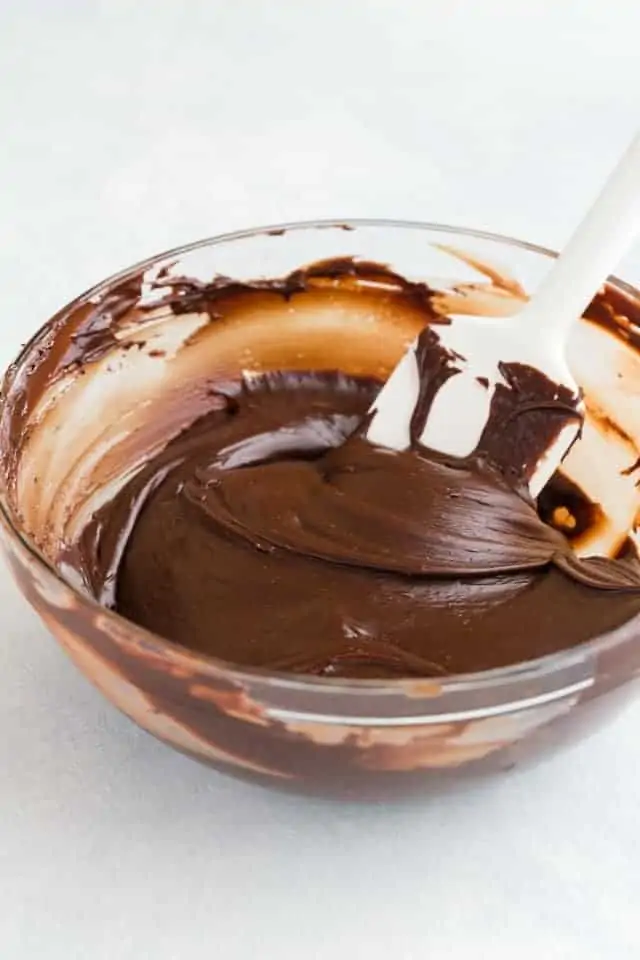 A clear mixing bowl filled with melted fudge that's being stirred with a white silicone spatula.