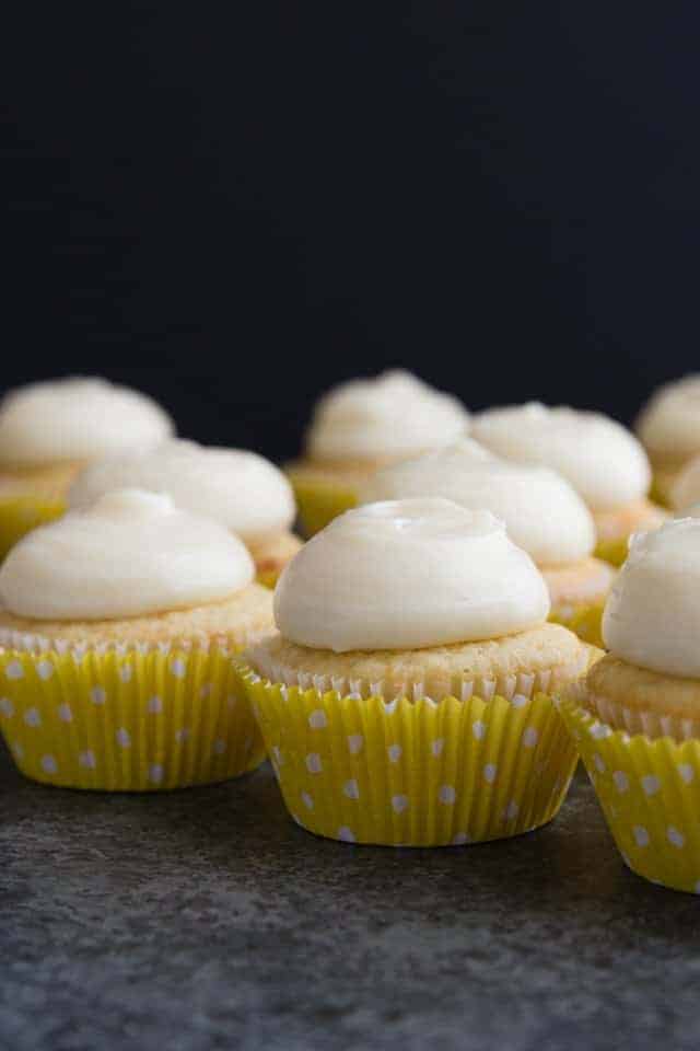 Vanilla cupcakes with lemon cream cheese frosting lined neatly on a dark tabletop.