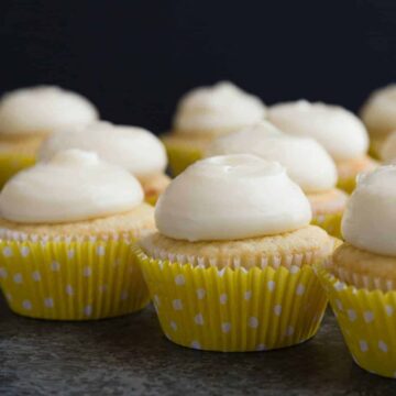 Vanilla cupcakes with lemon cream cheese frosting lined neatly on a dark tabletop.