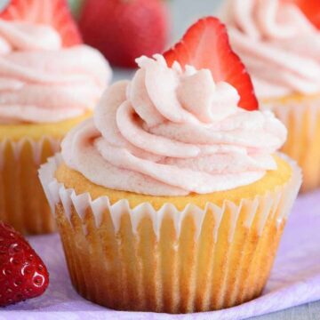 Vanilla yogurt cupcakes with strawberry frosting topped with a strawberry slice on a purple napkin.