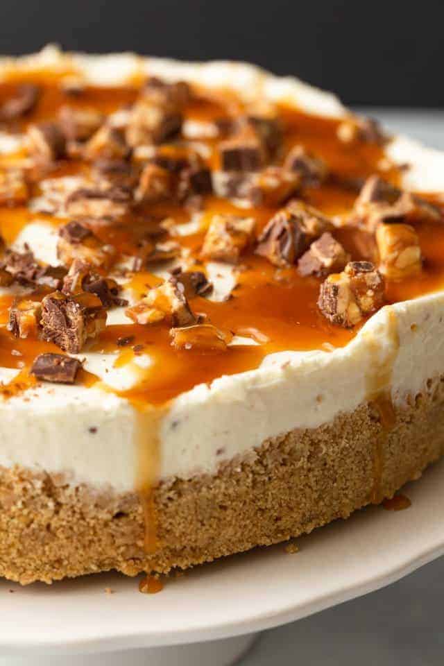 no bake snickers cheesecake with caramel drizzle on a white cake stand