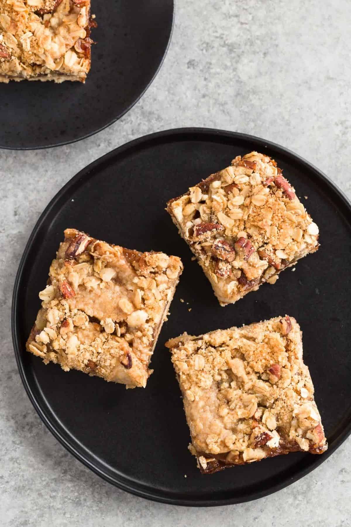 Three apple streusel bars on a black plate with a second bar, also on a black plate, sitting behind it.