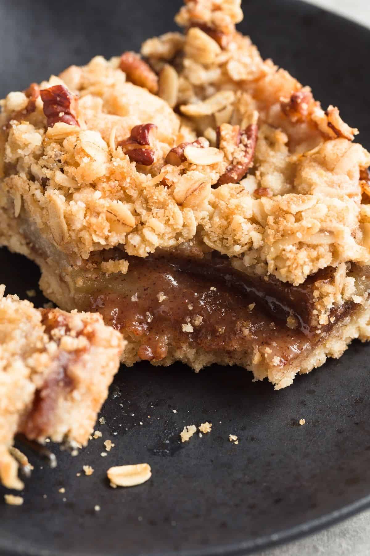 A close up view of an apple streusel bar on a black plate with a fork taking a bite out so the gooey apple filling is visible.