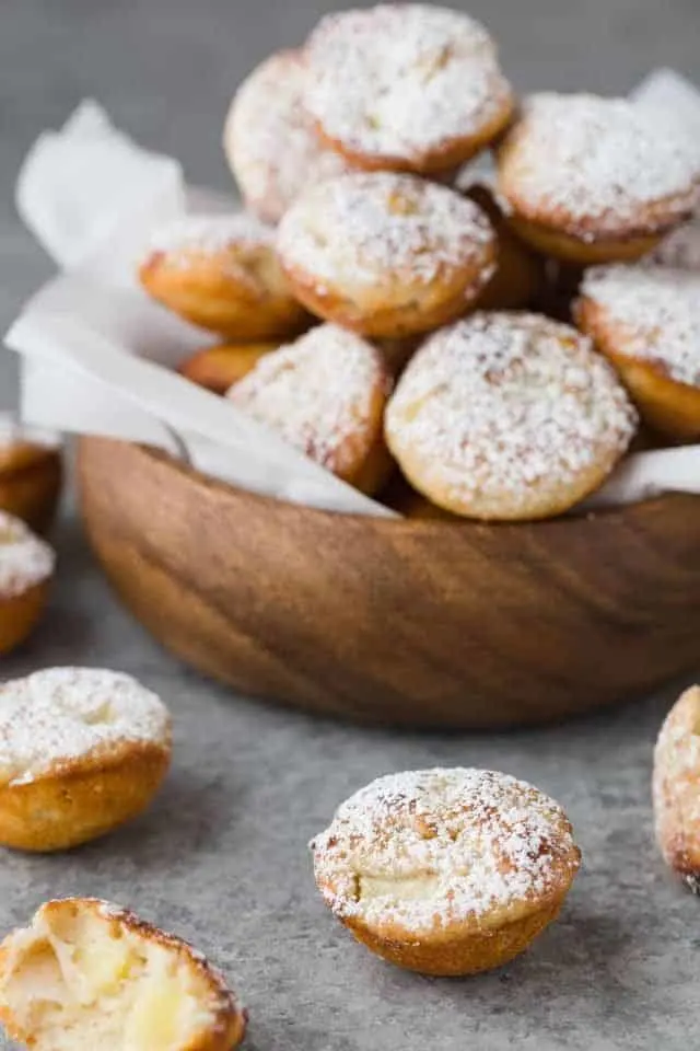 Baked apple fritters dusted with powdered sugar piled in a wooden bowl lined with white parchment paper and a few fritters scattered around the bowl.