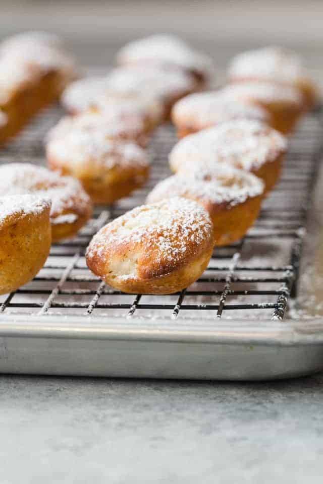 Side view of baked apple fritters dusted with powdered sugar lined up on a wire rack that is nestled in a stainless steel baking pan.