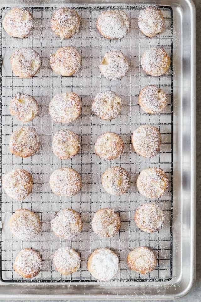 Overhead view of baked apple fritters dusted with powdered sugar lined up on a wire rack that is nestled in a stainless steel baking pan.