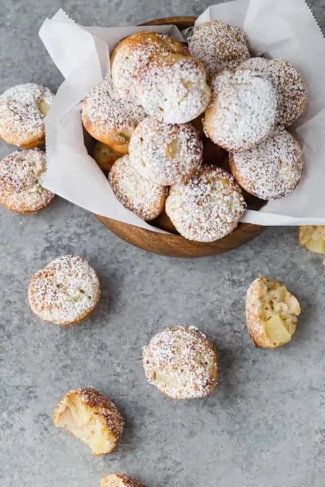 Overhead view of baked apple fritters dusted with powdered sugar piled in a wooden bowl lined with white parchment paper and a few fritters scattered around the bowl.