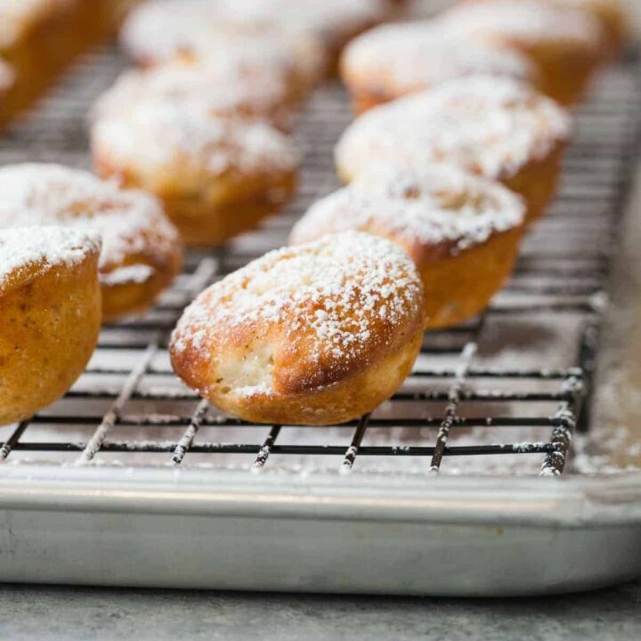 Side view of baked apple fritters dusted with powdered sugar lined up on a wire rack that is nestled in a stainless steel baking pan.