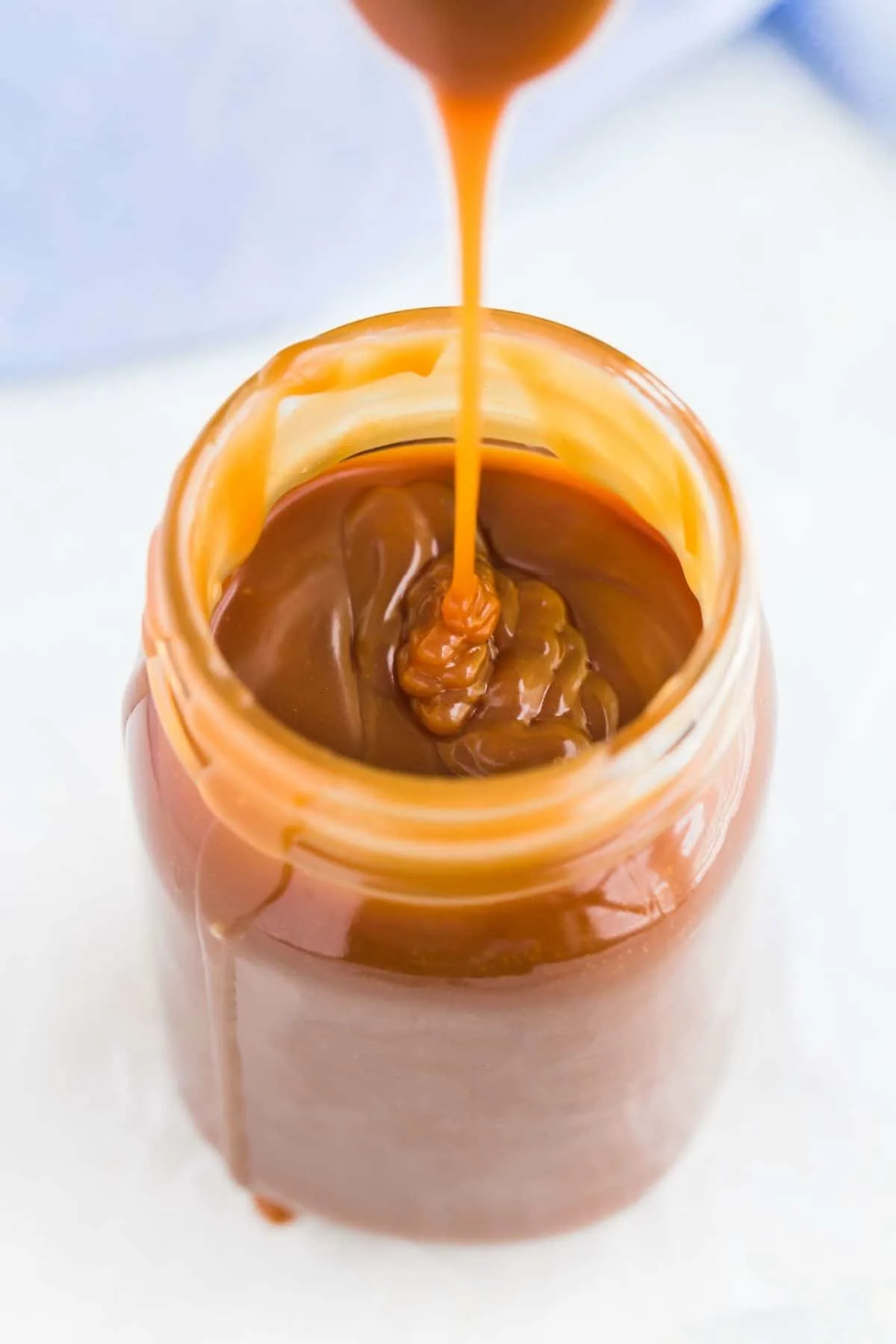 Homemade caramel sauce being drizzled in a tall mason jar with a light blue napkin behind it.