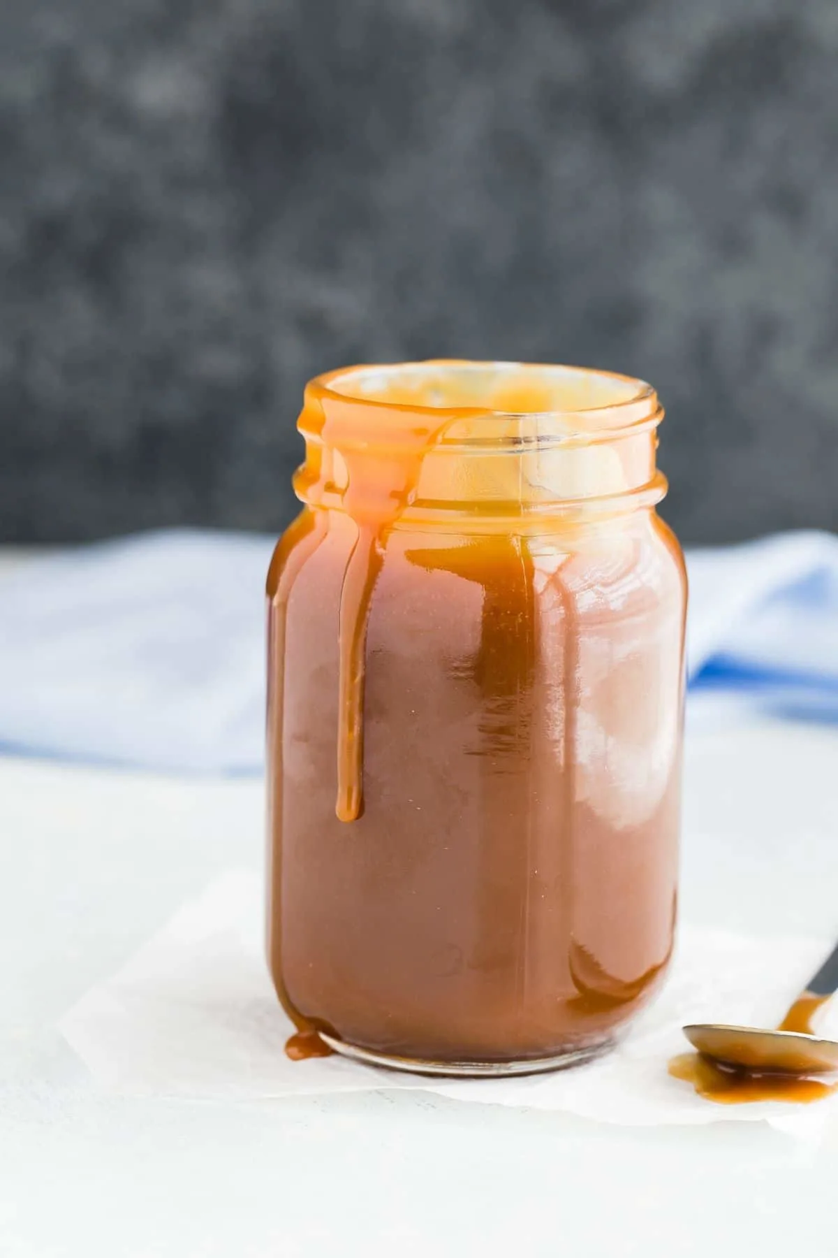 A jar of caramel sauce with caramel dripping down the side and a spoon sitting next to the jar.