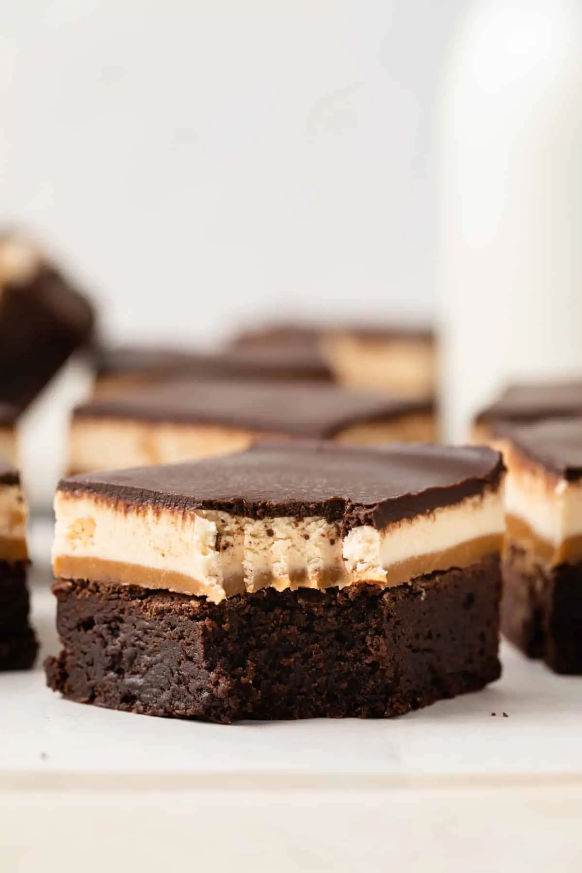 A fudgy chocolate brownie with layers of gooey caramel, chewy nougat, and chocolate ganache over the top sitting on white parchment paper with a fork taking a bite out.