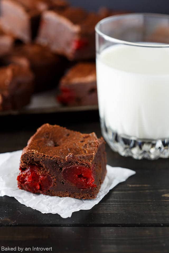 A single chocolate cherry brownie on a square piece of white parchment paper next to a glass of milk.