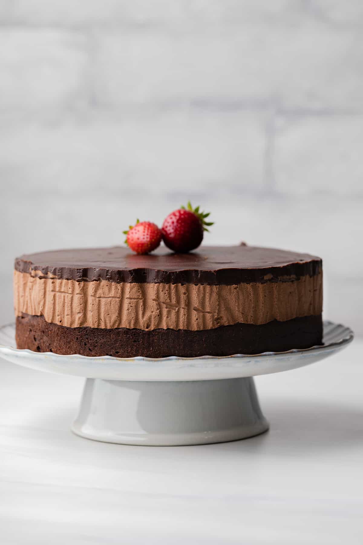 Triple chocolate mousse cake on a white cake stand with large strawberries stacked on top of the cake.