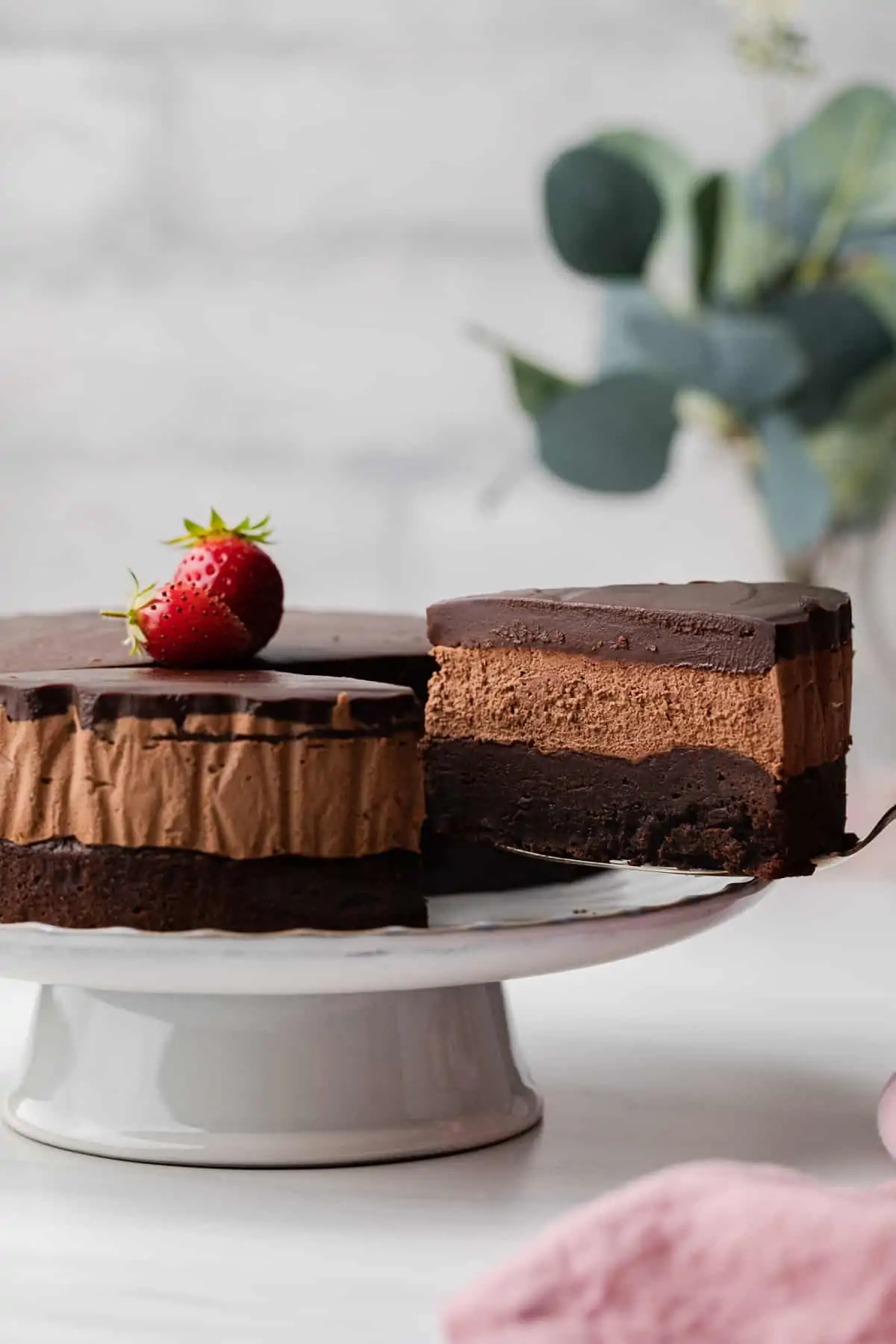 A close up view of triple chocolate mousse cake sitting on a white cake plate with large strawberries on top of the cake.