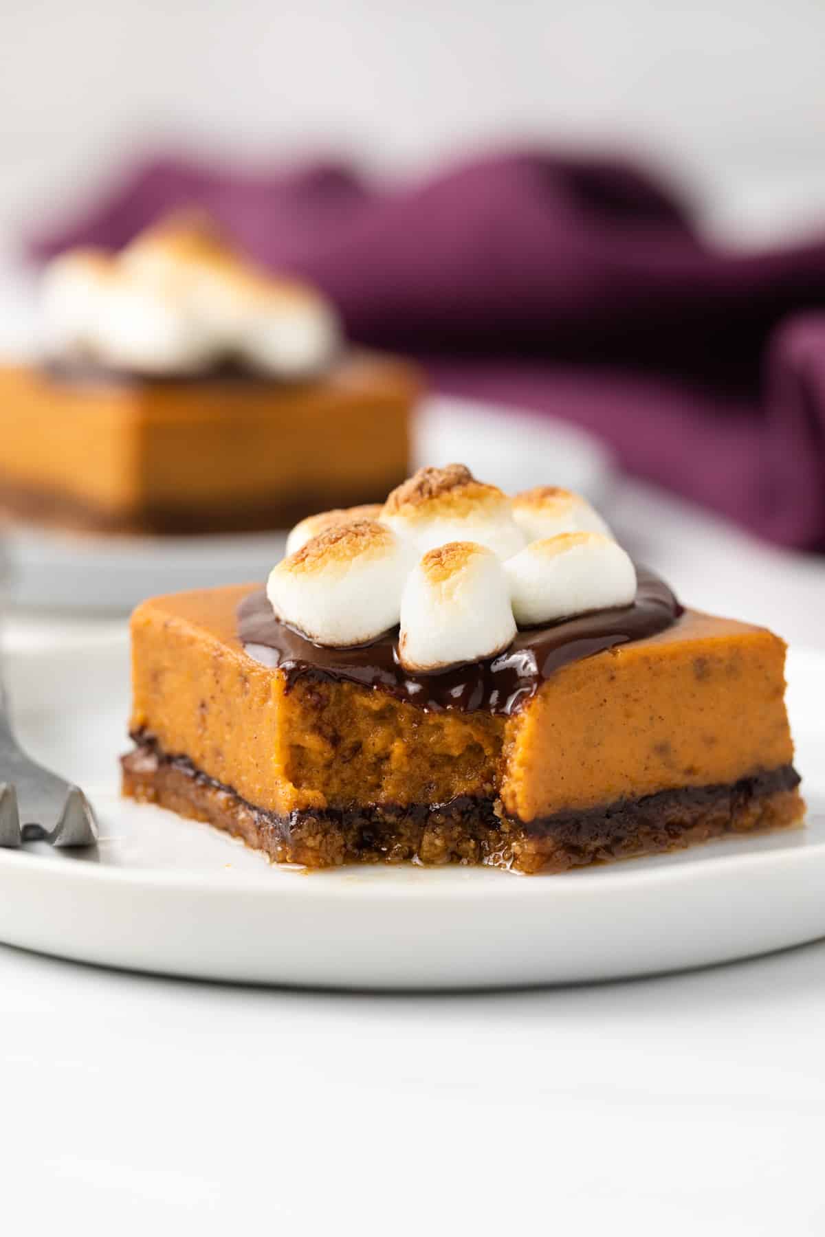 A pumpkin pie bar topped with chocolate ganache and toasted marshmallow on a white plate