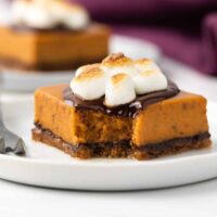 A pumpkin pie bar topped with chocolate ganache and toasted marshmallow on a white plate