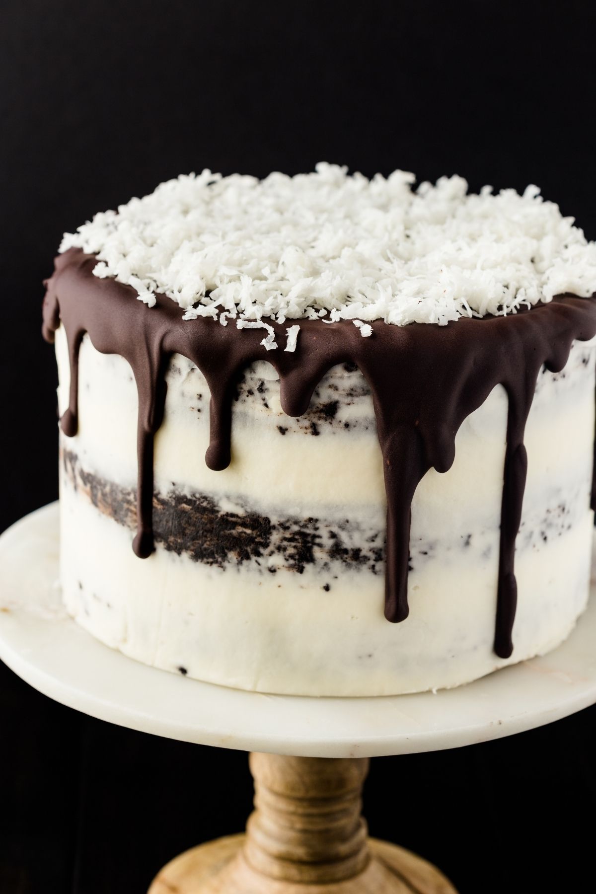 Chocolate coconut cake with dripping ganache and coconut flakes on top sitting on a cake stand.