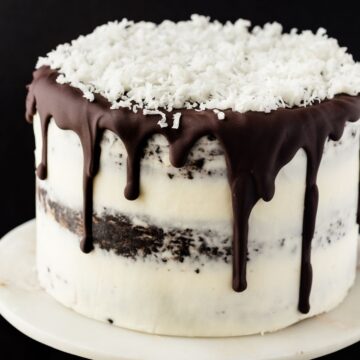 Chocolate coconut cake with dripping ganache and coconut flakes on top sitting on a cake stand.
