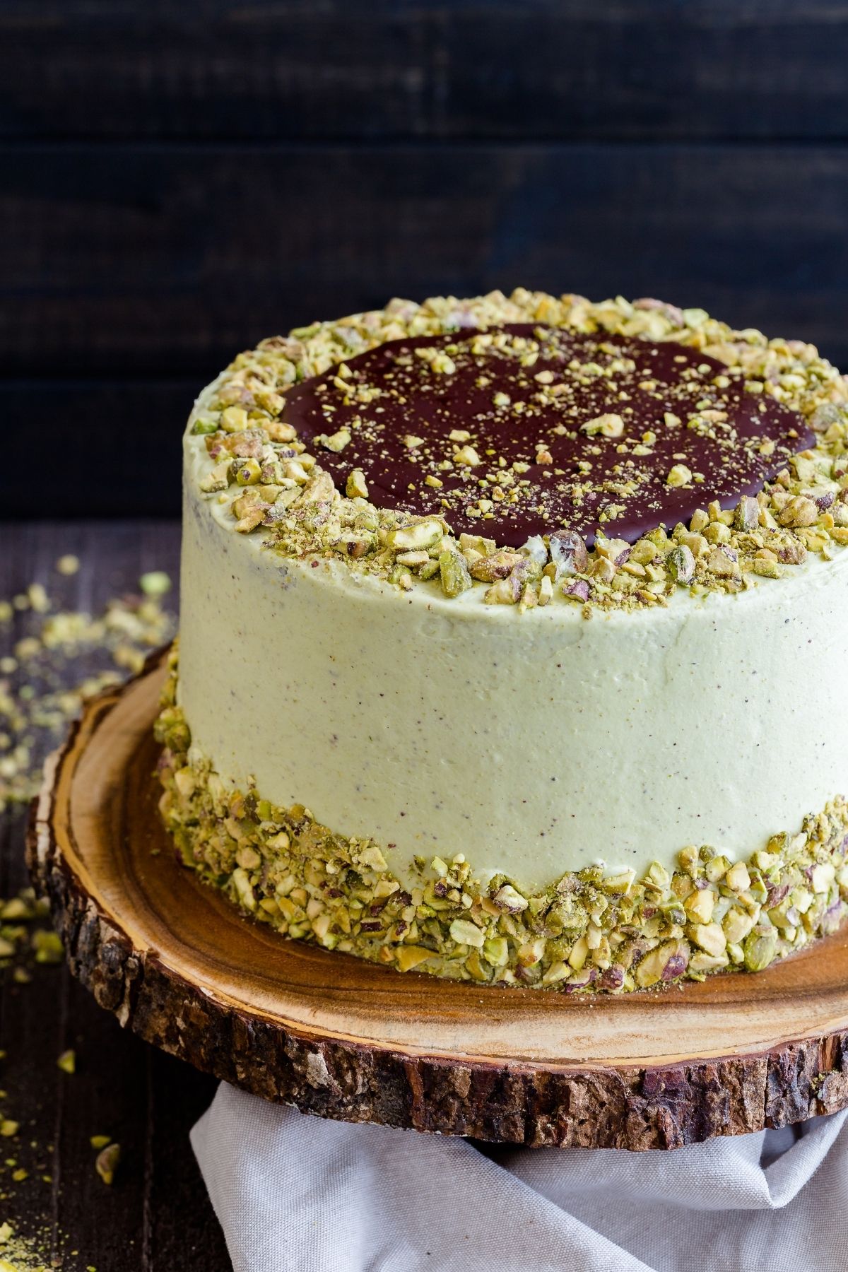 Chocolate pistachio cake on a wooden cake stand with a cream colored napkin around the base.