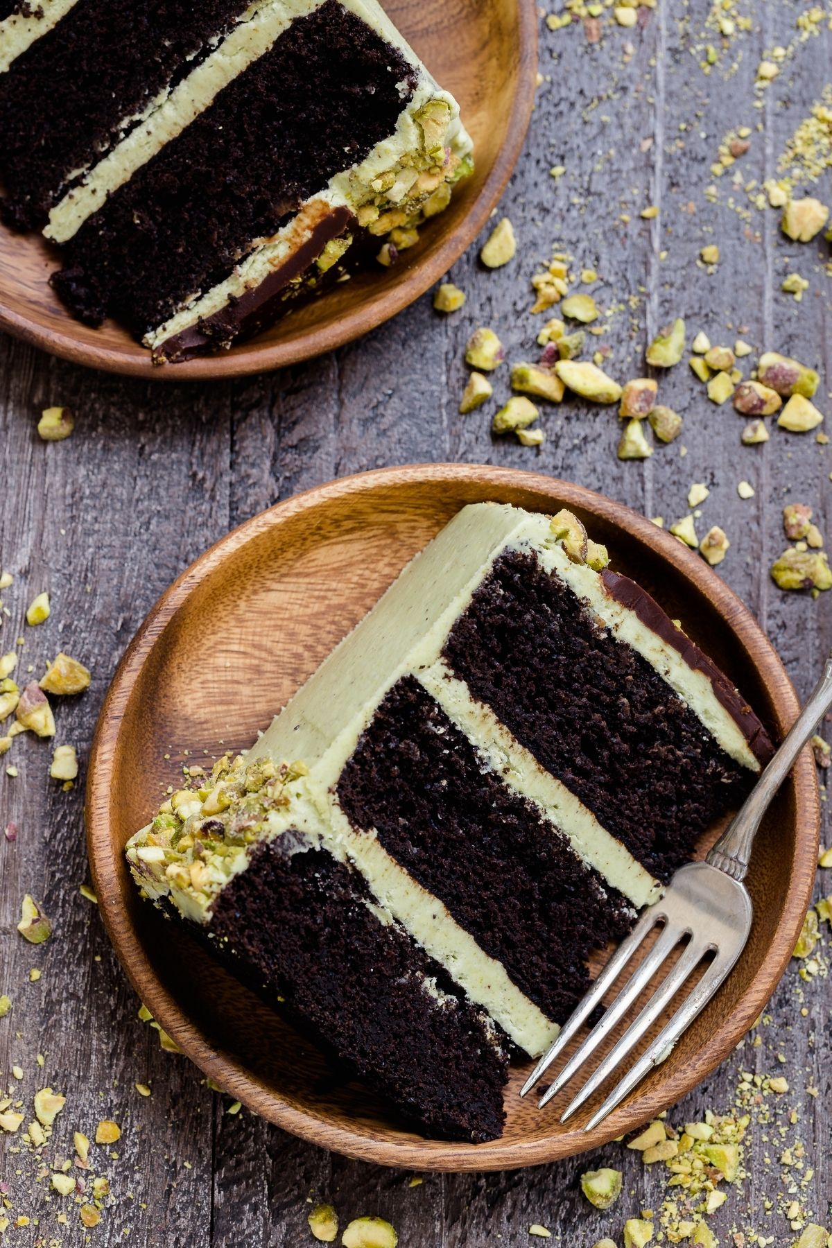 A slice of triple layer chocolate pistachio cake on a bamboo plate with a fork next to it.