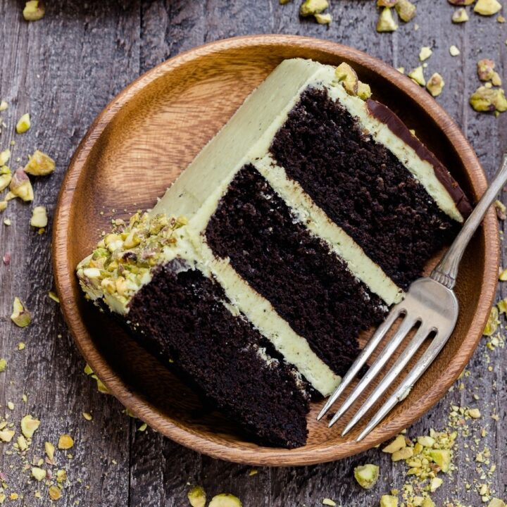 aA slice of triple layer chocolate pistachio cake on a bamboo plate with a fork next to it.