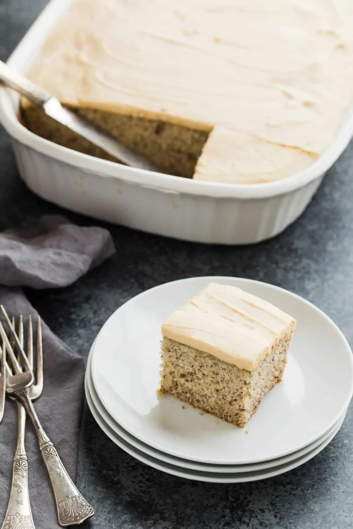 A slice of easy banana cake on a plate with a pan of banana cake behind it.