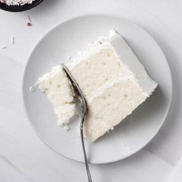 A slice of moist white cake with buttercream frosting on a plate, with a fork cutting off a bite