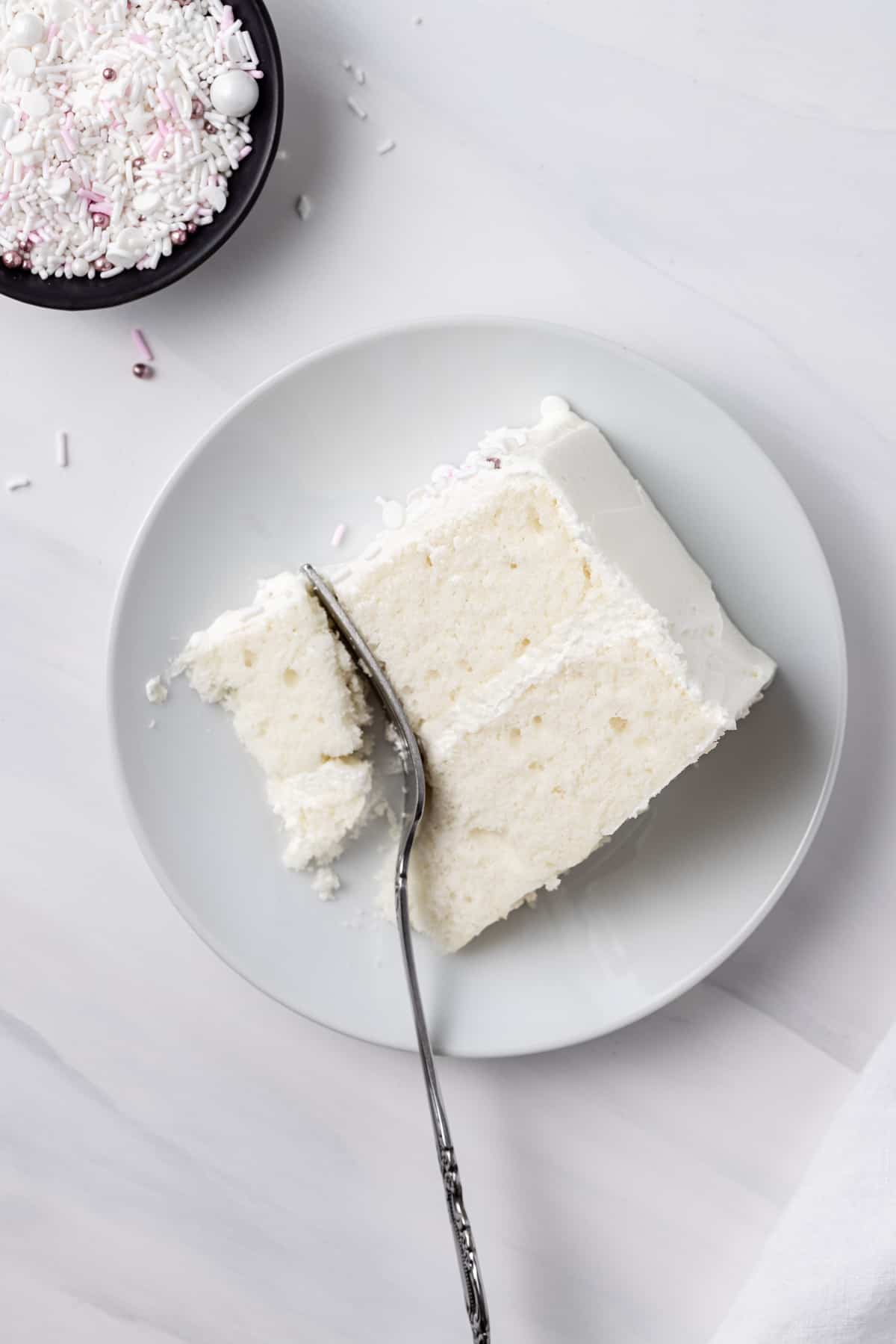 A slice of moist white cake with buttercream frosting on a plate, with a fork cutting off a bite