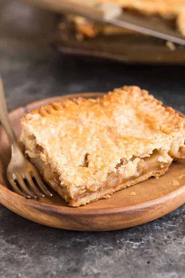 A square slice of apple slab pie on a wooden plate with a fork next to it.