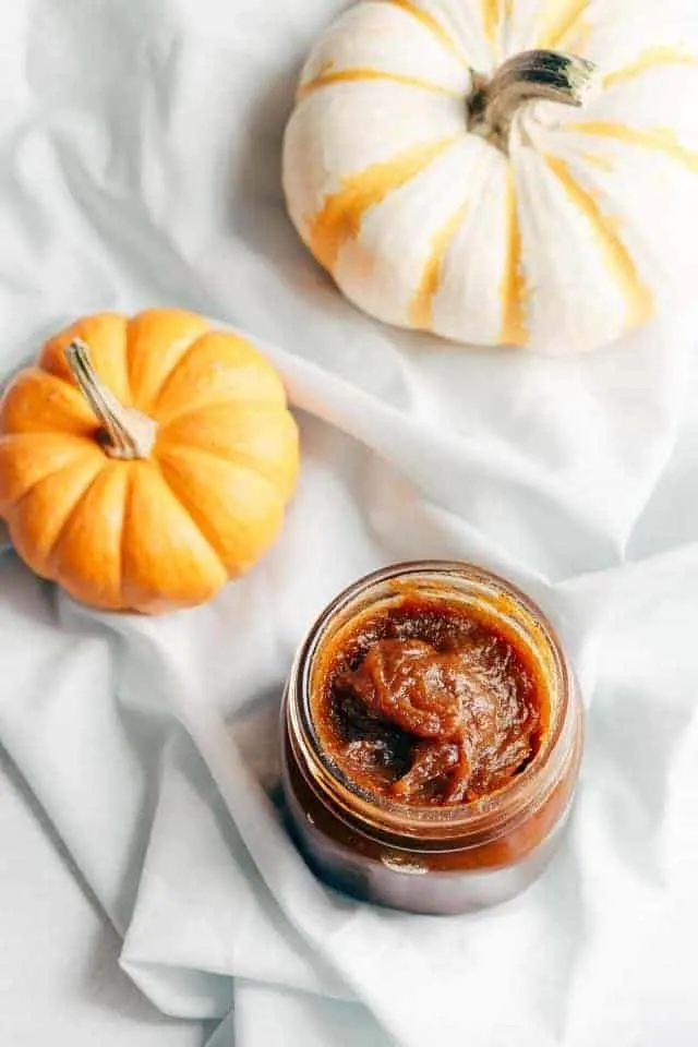 A glass jar of pumpkin jam, an orange pumpkin, and a white pumpkin sitting on a baby blue napkin.