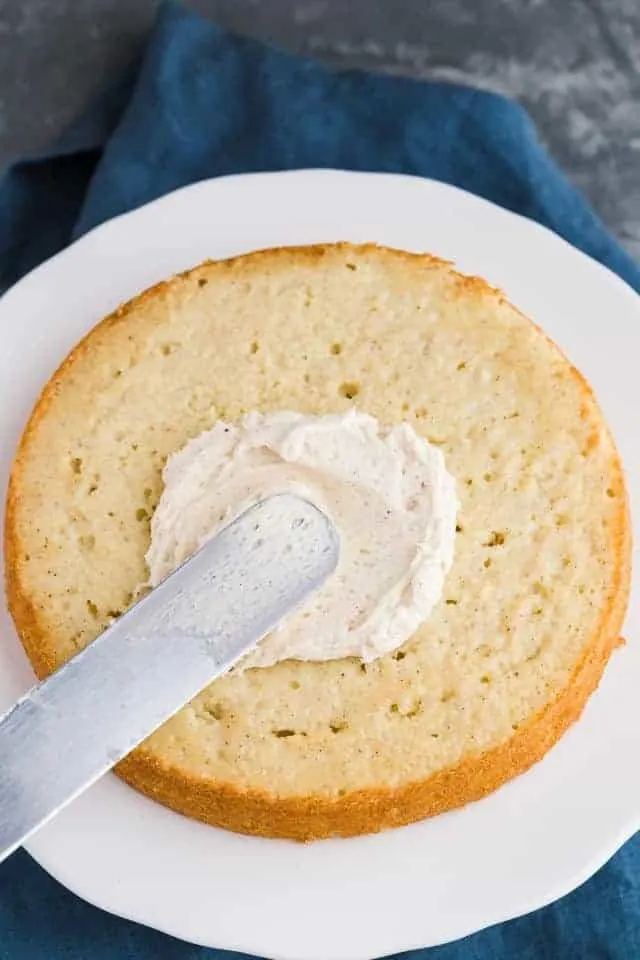 Frosting being spread over a layer of eggnog cake on a white cake stand.