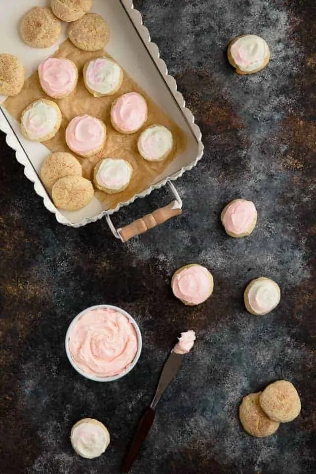 Frosted sour cream cookies next to a try filled with cookies and a white bowl filled with pink frosting.