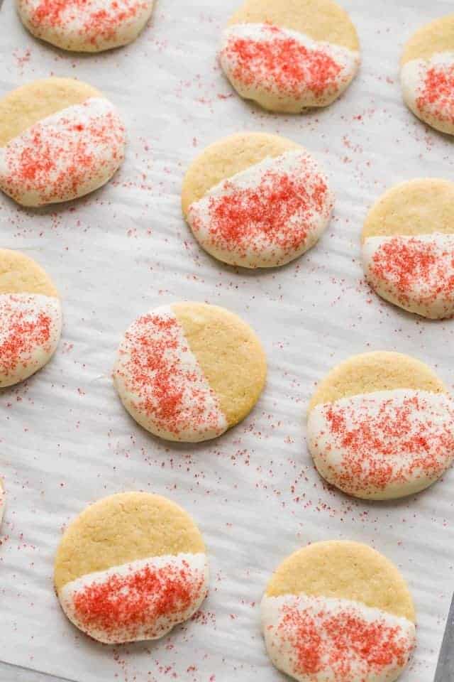 Peppermint cookies dipped in white chocolate and topped with red sanding sugar on a baking sheet line with white parchment paper.