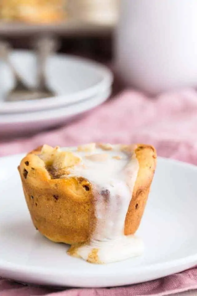 Close up view of Apple Pie Cinnamon Roll Cup on a white plate with glaze dripping down the side.