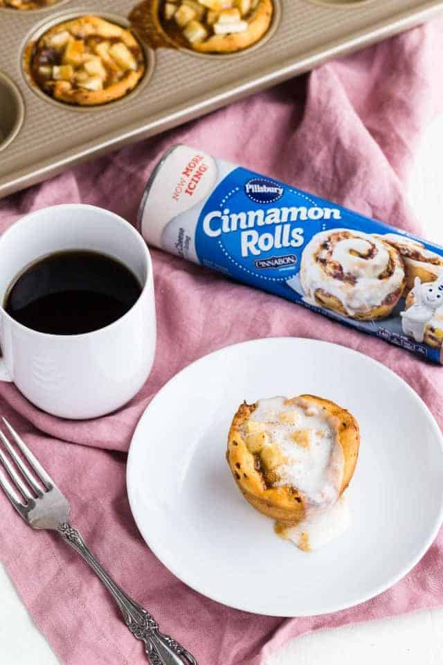 Apple Pie Cinnamon Roll Cup on a white plate with a fork and cup of coffee next to it.