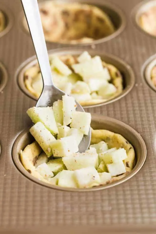 Cinnamon roll cups in a muffin tin being filled with chunks of apple.