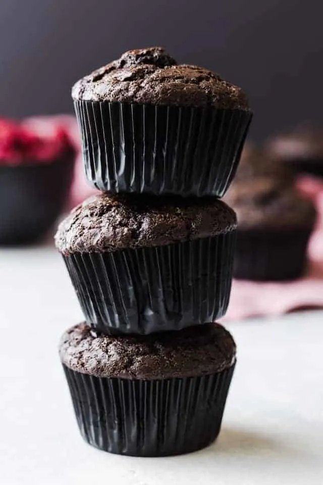 Three double chocolate muffins with raspberries stacked on a white tabletop.