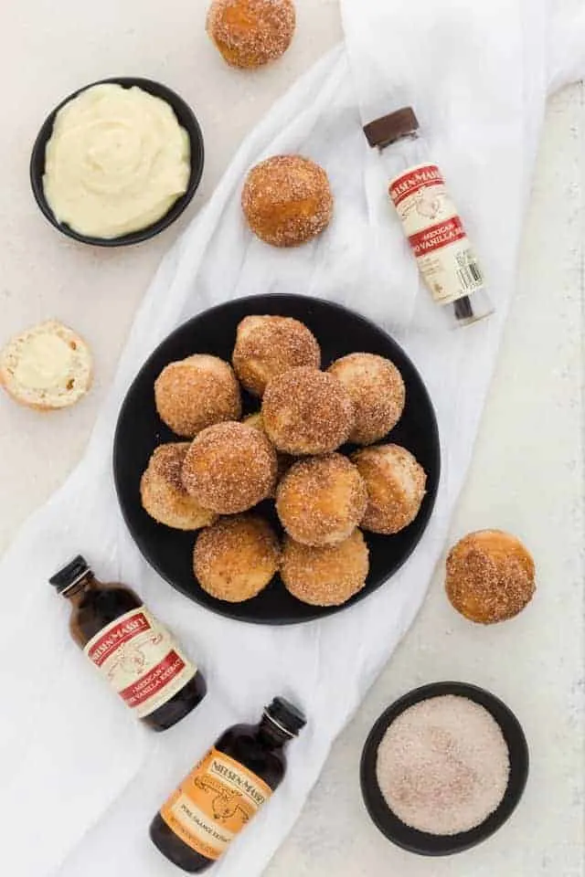 Overhead of baked bunuelos with anise custard on a black plate nest to Nielsen-Massey vanillas.