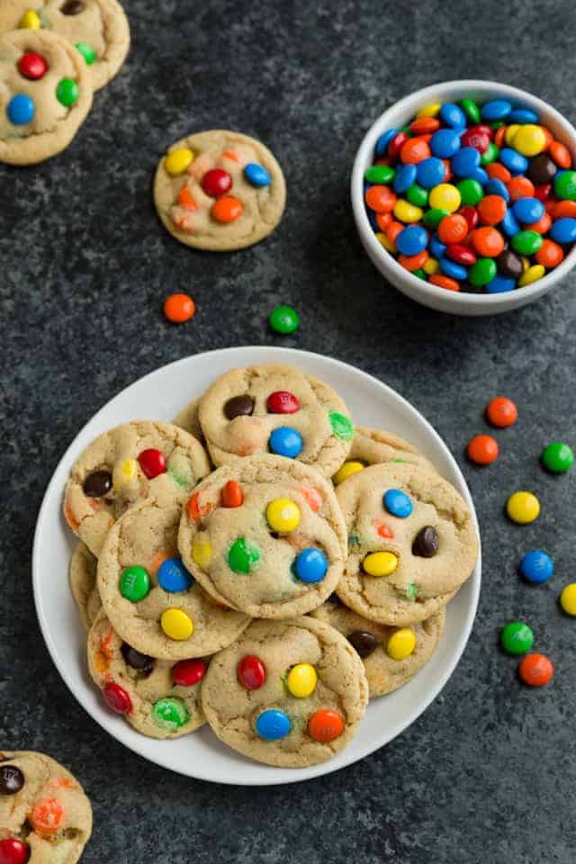 M&M cookies on a white plate with a bowl of m&m's next to it and a couple cookies scattered about on the table.