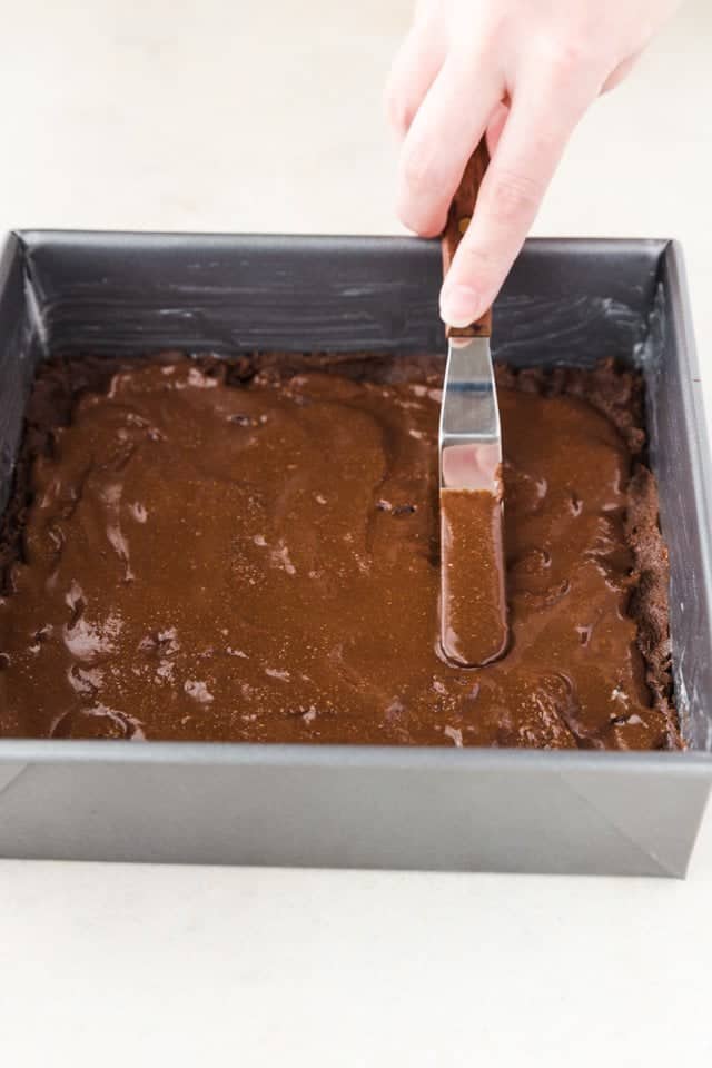 Nutella being spread over chocolate cookie dough in a square baking pan.