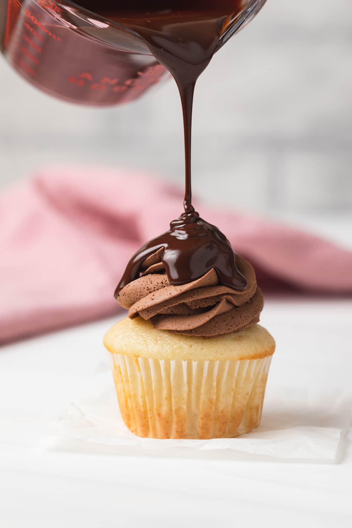 chocolate glaze being poured over chocolate frosting on a vanilla cupcake