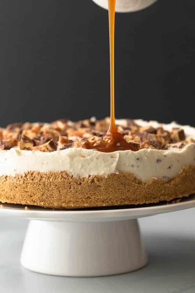 no bake snickers cheesecake with caramel drizzle on a white cake stand