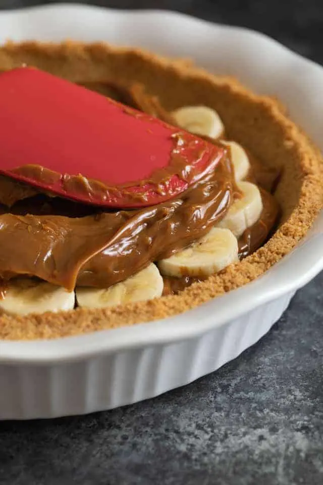 Dulce de leche being spread over sliced bananas in a graham cracker crust