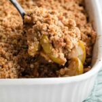 apple crisp being spooned out of a white casserole dish