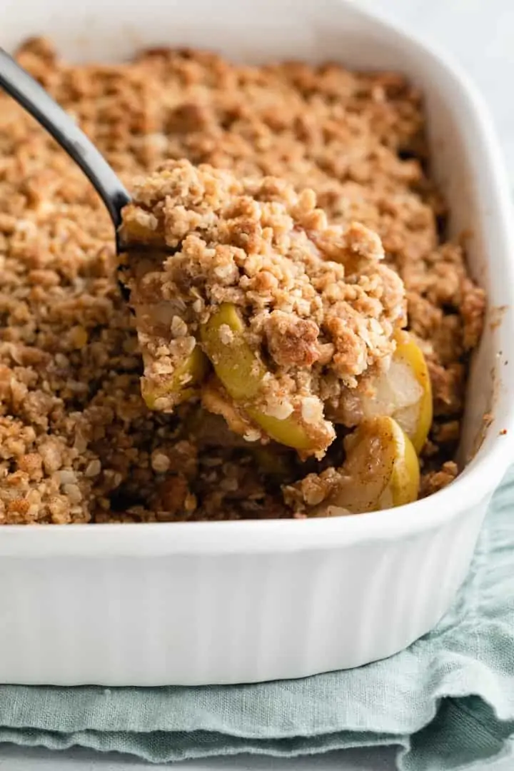 apple crisp being spooned out of a white casserole dish