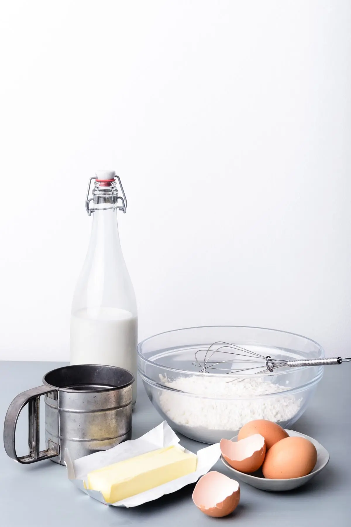 eggs, butter, flour in glass bowl, jar of milk, and metal flour sifter
