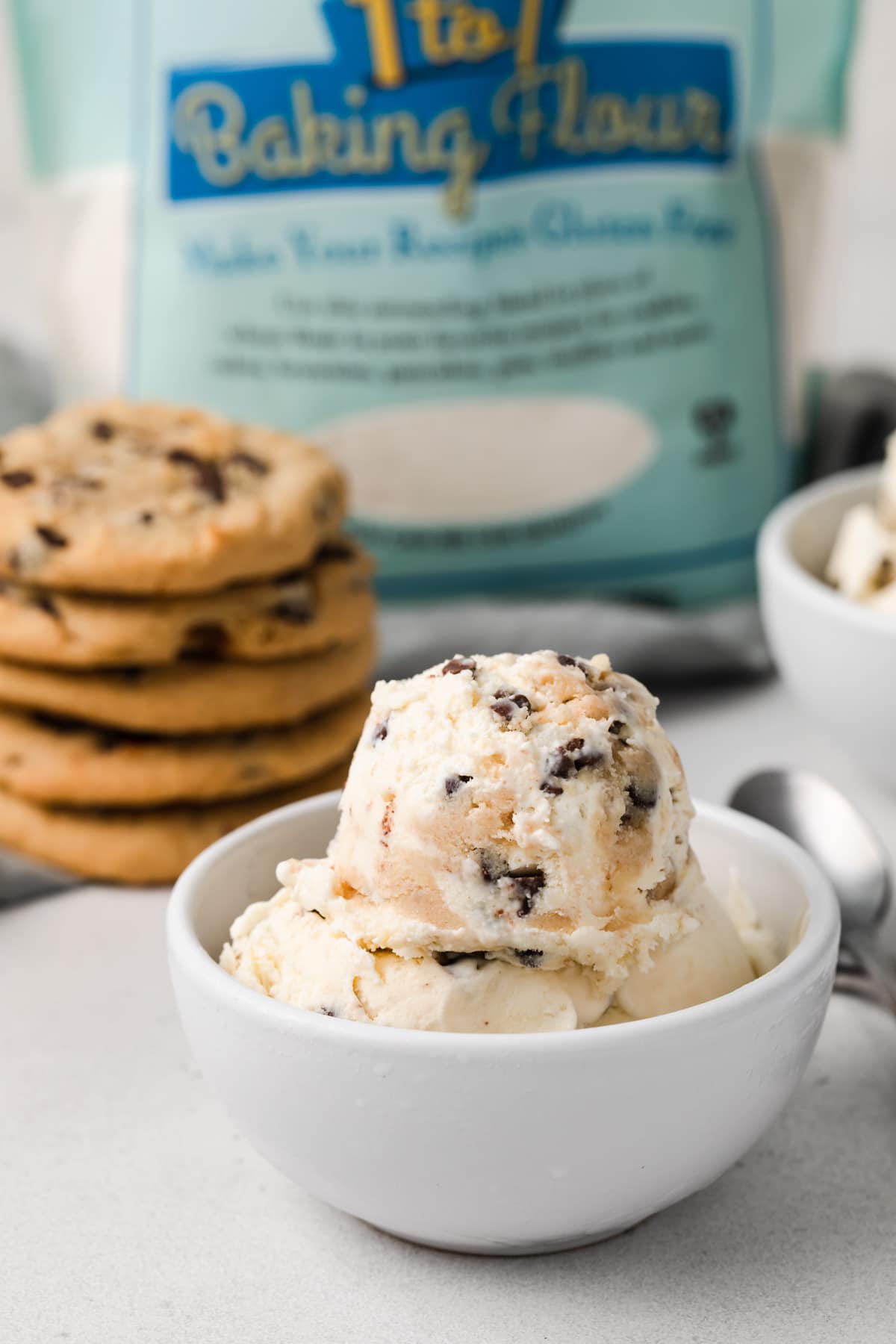 the best cookie dough ice cream scooped into white bowl with stack of chocolate chip cookies and bag of flour